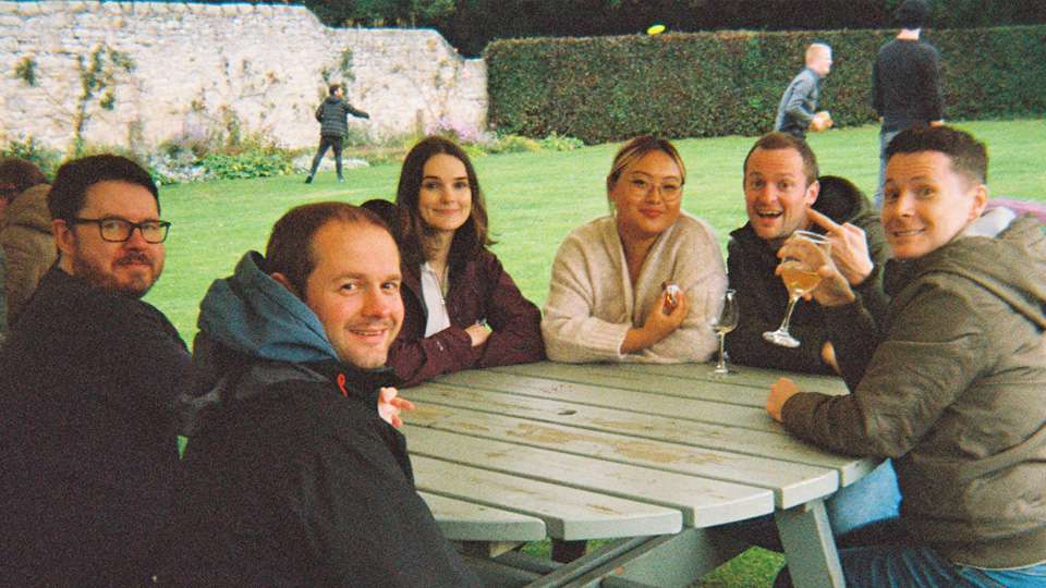Group of Storm employees socialising around a round picnic table in in a castle garden with sporting activities happening in the background
